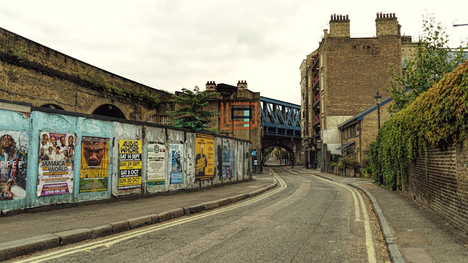 An empty urban street scene showing a gentle curve in the road accompanied by a double yellow line along the curb. On the left side, there is a low stone wall topped with a weathered wooden fence covered in colorful posters and advertisements, some promoting music events and local activities. Behind the fence, a red-brick building with arched openings and a small tree growing over the top is visible, with a blue metal bridge or viaduct crossing overhead. To the right, a tall brick wall with green ivy or foliage climbing up it runs alongside narrow pavement sections. The environment appears to be part of a residential or commercial area in Kentish Town, London, with an overcast sky providing diffuse lighting. The scene suggests a quiet, everyday street, potentially involved in home relocation or furniture transport activities by companies like Self Storage Kentish Town, as part of their removals and moving services offered in this area.