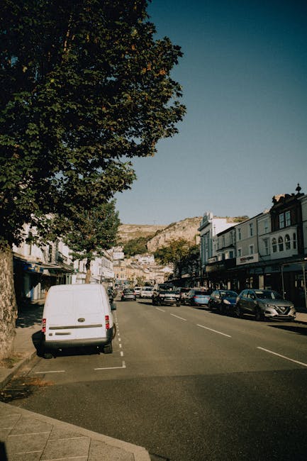 An aerial view of a residential area showing a busy intersection with multiple roads, houses with tiled roofs, and lined streets with parked cars. The image captures the intersecting roads crossing a railway line supported by concrete bridges, with green trees scattered along the streets and in yards. The vantage point reveals the layout of the neighbourhood, including driveway entrances, garden spaces, and the railway track structure. The detailed layout highlights the connection between transportation infrastructure and housing streets, illustrating a typical urban residential setting. This scene is relevant to house removals and moving services, such as those provided by Self Storage Kentish Town, as it depicts the environment where furniture transport, packing, and home relocation activities take place in a typical Kentish Town setting.