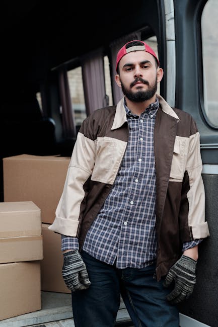 A man dressed in a blue jacket, navy trousers, and white sneakers is seen loading boxes into the rear of a white cargo van parked on a cobblestone street in an urban area, with multi-storey residentia
