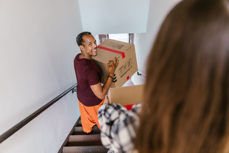 A man with short dark hair and a smiling expression, wearing a maroon t-shirt and orange trousers, is carrying a medium-sized cardboard box with red tape along the top, as part of a home relocation pr