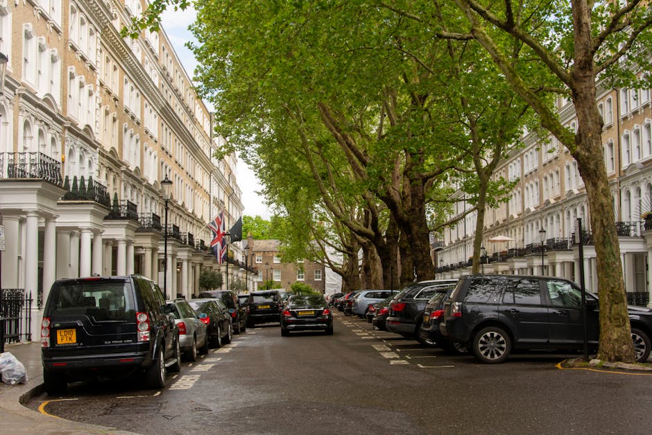 A residential street in Kentish Town with white Victorian-style terraced buildings featuring black wrought-iron balconies and decorative columns. Several parked cars, including a black van, an SUV, an